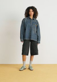 Veste en denim bleu délavé, coupe surdimensionnée avec une seule poche poitrine. Short cargo noir avec poches latérales. Baskets vert clair et chaussettes grises.