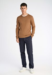 Maglione lavorato a maglia marrone, collo rotondo, abbinato a pantaloni blu scuro. Modella che indossa sneakers bianche, in piedi su uno sfondo semplice.