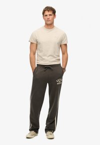 Lichtbeige T-shirt met korte mouwen en donkergrijze trainingsbroek met een wit logo. De broek heeft zijstrepen en wordt gedragen met beige sneakers.