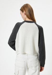 Häkelpullover mit cropped Schnitt, featuring einen weißen Körper und dunkelgraue Raglanärmel. Minimale Nähte und ein geripptes Kragenprofil.