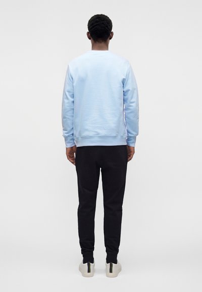 Homme debout de dos portant un sweat-shirt bleu clair, un pantalon noir et des baskets blanches avec des rayures vertes et jaunes.