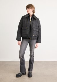 Casaco acolchoado preto com colarinho pontudo, ferragens douradas, dois bolsos frontais e fecho de correr, combinado com jeans cinzentos à boca de sino e botas pretas robustas.