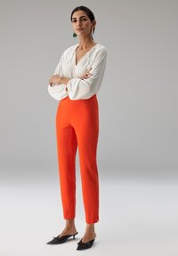 Witte blouse met geplooide mouwen gecombineerd met feloranje, broek met hoge taille. Zwarte mules met kleine strikjes aan de voorkant.