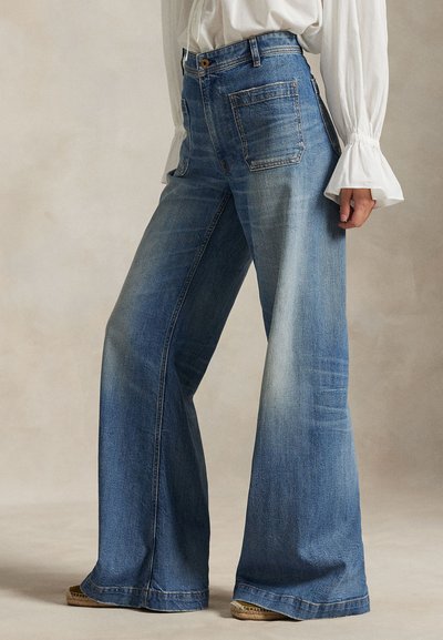 Femme portant un jean bleu taille haute à jambes larges avec des poches plaquées devant et un chemisier blanc à manches évasées.
