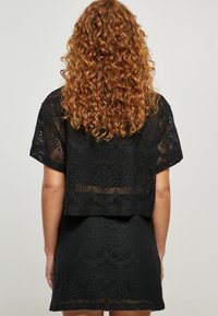 Schwarzes Häkel-Crop-Top, das über einem passenden Kleid getragen wird, mit einem strukturierten Muster und kurzen Ärmeln sowie einem offenen Rückendesign.