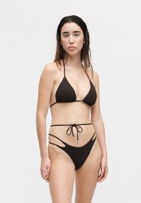 Mujer con el cabello mojado que lleva un bikini negro de tiras con detalles recortados, de pie contra un fondo blanco liso.