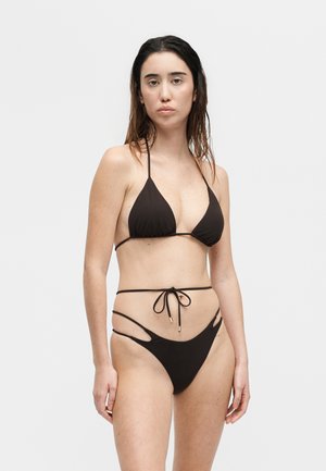 Γυναίκα με βρεγμένα μαλλιά που φοράει μαύρο στρινγκ μπικίνι με σχέδια κοπής, στέκεται μπροστά σε απλό λευκό φόντο.