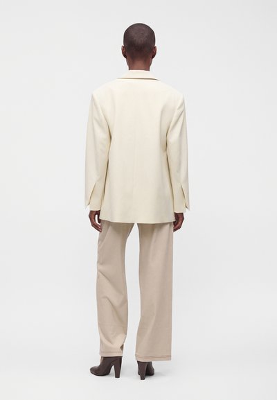 Personne portant un blazer crème, un pantalon beige à jambes larges et des chaussures à talons marron, debout de dos sur un fond blanc uni.