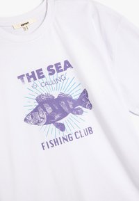 Witte t-shirt met paarse vissenillustratie en tekst "DE ZEE Roept, VISSENCLUB" op een achtergrond van paarse en blauwe stralen.
