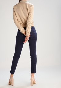 Béžová bomber bunda s elastickými manžetami a pasem, doplněná námořnickými slim-fit kalhotami a nahými vysokými sandály. Minimalistický design.