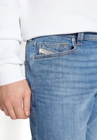 Hand resting beside light blue denim jeans pocket med synlig Diesel-etikett, iförd vit långärmad tröja.