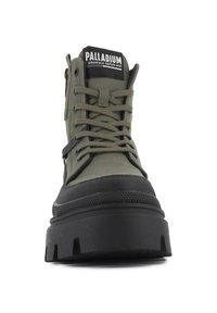 Botte montante vert olive avec semelle en caoutchouc noire, accents texturés, lacets à l'avant et fermeture éclair sur le côté. Présente le logo "PALLADIUM" sur la languette.