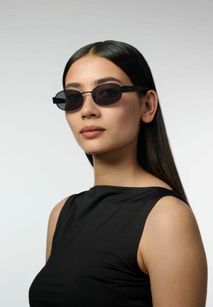 Femme aux longs cheveux foncés portant de petites lunettes ovales noires et un haut noir sans manches, légèrement tournée vers la droite sur un fond uni.