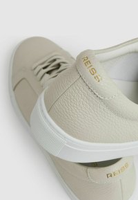 Adidași din piele de culoare bej deschis, cu o suprafață texturată, talpă albă și un branding subtil de aur pe călcâi. Design clasic cu șireturi.