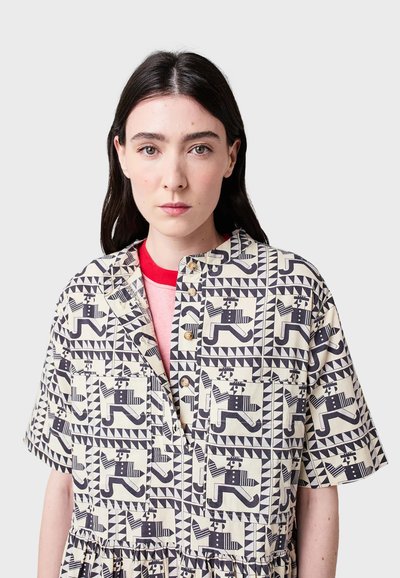 Giovane donna con lunghi capelli scuri che indossa una camicia a bottoni con motivo che presenta figure stilizzate di corridori neri su sfondo crema.