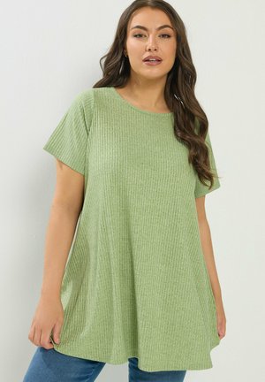 Femme aux longs cheveux bruns ondulés portant un haut vert clair côtelé à manches courtes et ample et un jean bleu sur un fond uni.