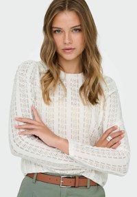 White lace sweater with long sleeves featuring intricate patterns and a round neckline, paired with a brown belt and casual pants.