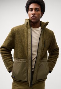 Grüne Fleecejacke mit einem weichen, strukturierten Finish, frontzip, zwei großen Taschen und einem kontrastierenden olivgrünen Stoff an der Taille und den Ärmeln.