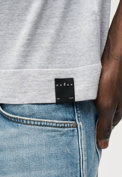 Κοντινό πλάνο ενός χεριού κοντά σε ανοιχτό μπλε τζιν και γκρι φούτερ με μαύρη ετικέτα που γράφει "Hogan Milano 1985."