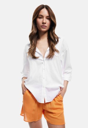 Hvid skjorte med knapper lavet af letvægtsstof, parret med løse orange shorts. Skjorten har oprullede ærmer og en afslappet pasform.