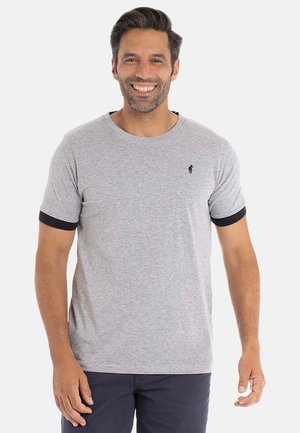 Homme souriant avec des cheveux noirs, portant un t-shirt gris avec des bords de manches noirs et un pantalon bleu marine, se tenant contre un fond blanc.
