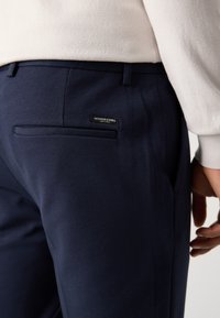Marineblauwe broek van een gestructureerde stof. Beschikt over een achterzak met een label. De zijaanzicht toont een soepele pasvorm en een gesneden design.