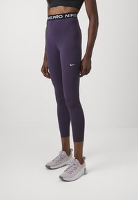 Leggings Nike Pro púrpuras con una cinturilla alta que presenta un logo negro. Combinados con zapatillas deportivas de color púrpura claro, creando un look deportivo y ajustado.