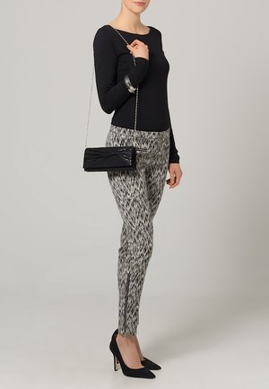 Mujer con top negro, pantalones grises con estampado y cremalleras, tacones negros puntiagudos y llevando un bolso pequeño negro con correa de cadena.