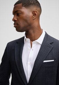 Marineblauwe blazer met een subtiel ruitpatroon, notch-lapels, een witte pochette en een witte overhemd met kraag. Glad stoftextuur.