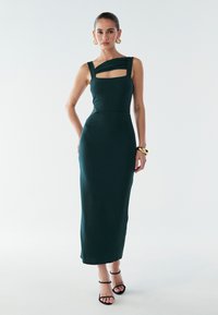 Ciemnozielona dopasowana sukienka z kwadratowym dekoltem, asymetrycznym paskiem na ramię i długością midi. Stylizowana z czarnymi sandałami na obcasie.