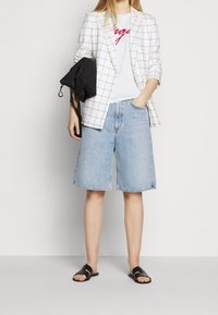 Blazer blanc avec un motif à carreaux noirs, t-shirt blanc avec un texte rouge, shorts en denim bleu clair et sandales noires à glissière, créant un look superposé.