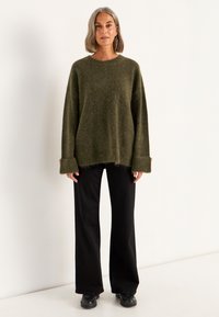 Olijfgroene gestructureerde trui met een losse fit, verlaagde schouders en brede manchetten, gecombineerd met zwarte culottes en zwarte schoenen.