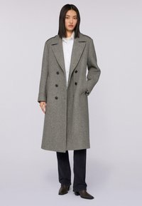 Šedý dlouhý dvouřadý kabát s širokými klopami, vyrobený z texturované tkaniny. Obsahuje černé knoflíky a boční kapsy, nasazený přes bílou košili.
