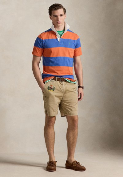 Πόλο με κοντά μανίκια σε μπλε και πορτοκαλί οριζόντιες ρίγες, φορεμένο πάνω από ένα πράσινο ριγέ μπλουζάκι. Μπεζ σορτς με κεντημένες λεπτομέρειες, καφέ παπούτσια σκάφους.