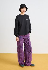 Chemise noire à manches longues, pantalon cargo violet avec plusieurs poches, et un bob noir. Porte des chaussures vertes. Fond uni.