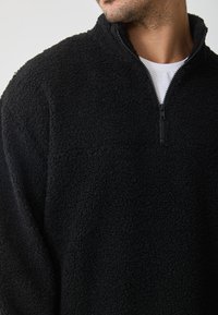 Pullover in pile nero con colletto alto e design a mezza zip. Tessuto testurizzato con una sensazione morbida e vaporosa. Nessun logo o accento visibile.