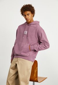 Sudadera de color púrpura claro con bolsillo frontal tipo canguro y capucha con cordón. Presenta un parche con el logo en el pecho. Combinada con pantalones beige.