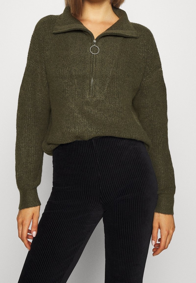 Femme portant un pull en maille vert olive avec une fermeture éclair sur le devant et un pantalon en velours côtelé noir, visible du cou jusqu'aux cuisses.