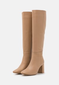 Paire de bottes en cuir beige montant jusqu'au genou avec talons carrés et fermeture éclair latérale, posées debout sur un fond blanc uni.