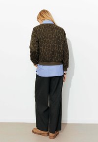 Veste bomber vert olive avec imprimé léopard, poignets et ourlet côtelés ; superposée sur une chemise bleu clair boutonnée et un pantalon large foncé.