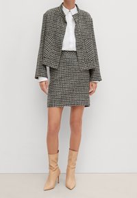 Veste en tweed noir et blanc avec poches avant, assortie à une jupe coordonnée. Des bottes à talons hauts beige complètent la tenue.