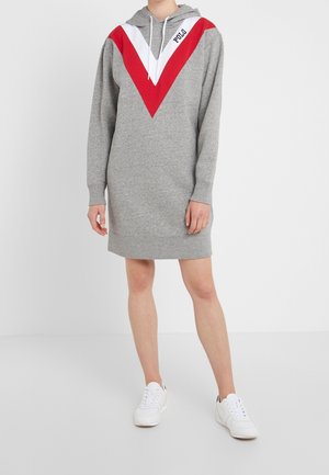 Personne portant une robe sweat à capuche grise avec un motif chevron rouge et blanc et des baskets blanches, debout devant un fond blanc uni.