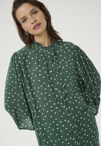 Blouse verte à pois avec un col froncé, des manches bouffantes longues et des boutons bleus. Le tissu a une apparence texturée.
