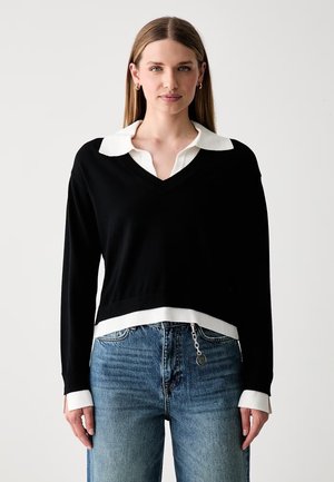 Donna con capelli lisci che indossa un maglione nero a scollo a V sopra una camicia bianca con colletto e jeans blu con un accessorio a catena.