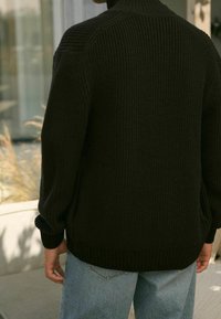 Ciemnozielony sweter wykonany na drutach z wysokim kołnierzem, prążkowaną teksturą i luźnymi rękawami, w połączeniu z jasnoniebieskimi dżinsami.
