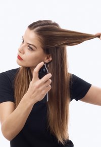 Femme en chemise noire pulvérise un produit capillaire sur une section de cheveux bruns longs maintenue tendue, se préparant à la coiffer.
