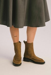 Brązowe zamszowe botki za kostkę z płaską czarną podeszwą, z bocznymi szwami i wyższym stanem nad kostką, w połączeniu z szarym spódnicą.