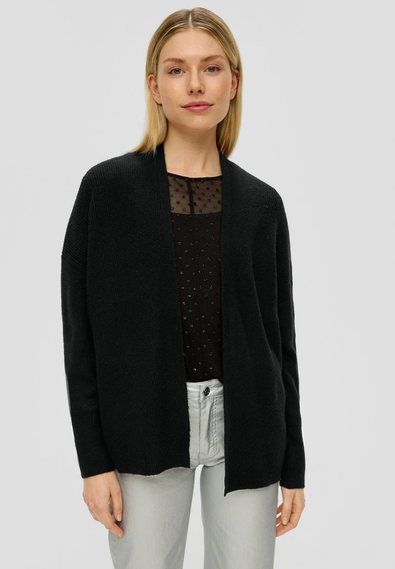 Schwarze Strickjacke mit offenem Vorderteil, geripptem Muster und langen Ärmeln, kombiniert mit einem schwarzen durchsichtigen Oberteil mit kleinen gestickten Punkten.