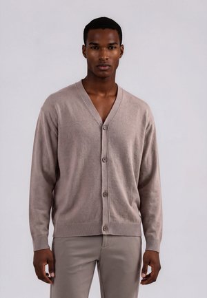 Jeune homme portant un cardigan beige boutonné et un pantalon assorti, debout devant un fond gris clair.