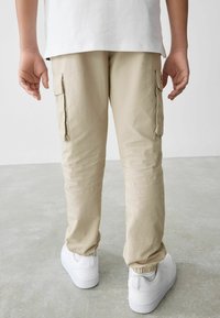 Beige cargo-housut, joissa on sivutaskut ja elastiset hihnat, yhdistettynä valkoisiin lenkkikenkiin. Materiaali vaikuttaa kevyeltä ja rakenteelliselta.